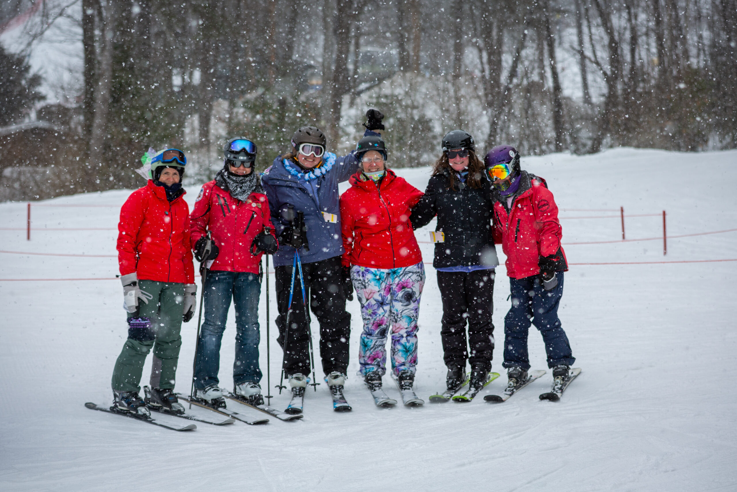 Six skiers in colorful jackets pose together on a snowy slope as snow falls, with trees in the background.